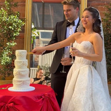 Load image into Gallery viewer, Couple cutting a wedding cake with a red tablecloth and mirror reflection.