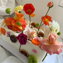Load image into Gallery viewer, A rectangular white cake decorated with a dense arrangement of colorful edible flowers, including orange, red, pink, purple, yellow, and white blooms, with dried citrus slices on top.