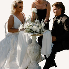 Load image into Gallery viewer, Wedding couple sitting outdoors with a bridesmaid, surrounded by greenery and a body of water.