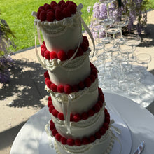 Load image into Gallery viewer, A white tiered wedding cake decorated with red raspberries and white ribbons, displayed outdoors on a white table with champagne glasses stacked in the background.