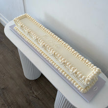Load image into Gallery viewer, A long rectangular cake decorated with cream-colored frosting and piped borders, displayed on a white pedestal table in a clean indoor setting.