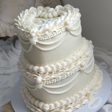 Load image into Gallery viewer, A three-tiered white cake decorated with intricate piped frosting details, draped ribbons, and shell motifs, displayed on a white board against a draped fabric background.