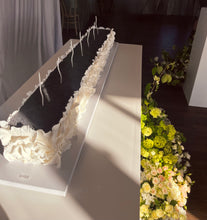 Load image into Gallery viewer, A long rectangular bridal cake with a black poppy seed center and white ruffled fondant sides, decorated with white wavy candles, displayed on a white table in a wedding venue setting.