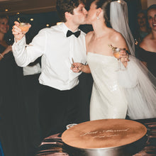 Load image into Gallery viewer, Couple kissing at a wedding reception with a cake in the foreground.