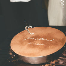 Load image into Gallery viewer, Round wedding cake with 'Just Married' inscription, couple in background holding drinks.