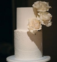 Load image into Gallery viewer, A two-tier cake with a stucco finish on the bottom tier and hand-carved flutes on the top tier, adorned with three fresh roses on the side.