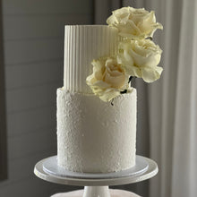 Load image into Gallery viewer, A two-tiered round cake with a textured stucco finish on the bottom tier and hand-carved flutes on the top tier, decorated with fresh roses, displayed on a white stand.

