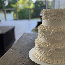Load image into Gallery viewer, Three-tiered white wedding cake on a wooden table with a blurred outdoor background
