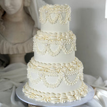 Load image into Gallery viewer, Three-tiered white wedding cake with decorative piping on a white ganache base.
