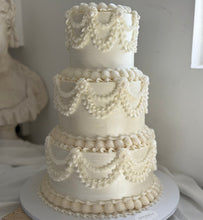 Load image into Gallery viewer, Three-tiered white wedding cake with decorative elements on a white surface.
