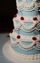 Load image into Gallery viewer, A close-up of a tiered cake with light blue frosting and white piped ruffle borders, decorated with red cherries on a white board against a dark background.
