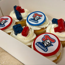 Load image into Gallery viewer, A box of frosted cupcakes decorated with white icing and round team toppers featuring a knight logo, along with red and blue candy bricks, shown inside a white box.
