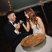 Load image into Gallery viewer, Bride and groom smiling while feeding each other tiramisu wedding cake at an evening wedding reception under festoon lights.
