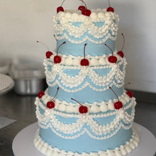 Load image into Gallery viewer, A three-tiered cake decorated with light blue frosting and white piped scalloped borders, topped with red cherries, displayed on a white round board.
