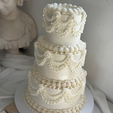 Load image into Gallery viewer, A three-tiered white cake decorated with intricate piped cream-colored frosting patterns, displayed on a white cake stand against a draped white fabric background with a classical bust statue in the background.
