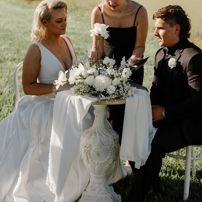 Wedding couple sitting outdoors with a bridesmaid, surrounded by greenery and a body of water.