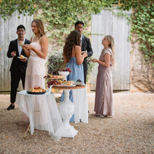 Load image into Gallery viewer, Group of people gathered around a table with food outdoors under greenery
