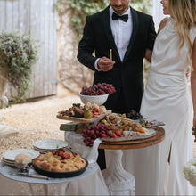 Load image into Gallery viewer, Man in tuxedo and woman in wedding dress standing near a table with food outdoors.
