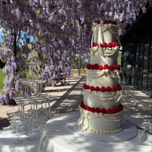 Load image into Gallery viewer, A tall white tiered wedding cake decorated with red berry-like accents and white draping, displayed on a white-draped table outdoors under a canopy of hanging purple wisteria flowers. Champagne flutes are arranged nearby, with a garden and building visible in the background.
