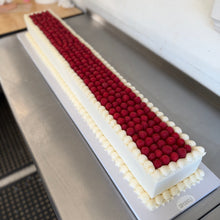 Load image into Gallery viewer, A long rectangular frosted cake or dessert display on a metal counter, decorated with a white border and a central strip of dark red raspberries.
