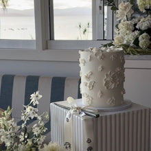 Load image into Gallery viewer, White wedding cake with floral decorations on a table with flowers around, by a window.
