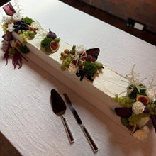 Load image into Gallery viewer, Long rectangular cake with floral decorations on a white tablecloth, accompanied by a knife and spatula.

