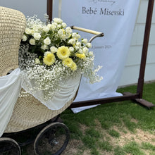 Load image into Gallery viewer, Wicker baby carriage with flowers in front of a banner with text
