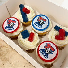 Load image into Gallery viewer, Six frosted cupcakes arranged inside a white cardboard box. Each cupcake has white icing and a round topper featuring a knights logo in blue on a red-and-white background and chocolate lego.
