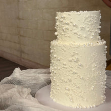 Load image into Gallery viewer, A two-tier white wedding cake decorated with white pearl-like beads and textured frosting, displayed on a white cake stand with draped tulle fabric in the foreground.
