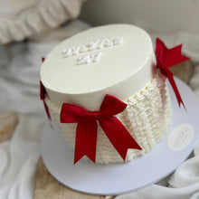Load image into Gallery viewer, A round white frosted cake with textured piped detailing like a cable knit sweater, red ribbon bows, displayed on a white round board with draped fabric in the background.

