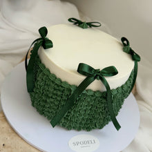 Load image into Gallery viewer, A round cake with a smooth off-white top and dark green ruffled piped sides like a christmas sweater, decorated with matching dark green ribbon bows, displayed on a white round board labeled 'SPODELI'.
