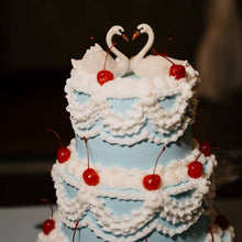 Load image into Gallery viewer, A three-tiered wedding cake decorated with light blue frosting, white piped ruffles, red cherries, and two white swan toppers on top that form a heart shape.

