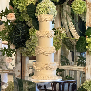 A four-tier white wedding cake with decorative details and green hydrangea flowers on each tier, displayed in a floral setting.