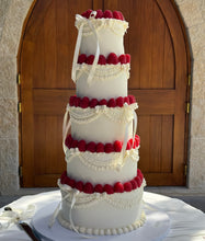 Load image into Gallery viewer, A five-tier white wedding cake decorated with red raspberries, white piped borders, and white ribbon bows, displayed on a white tablecloth in front of a large wooden arched door.
